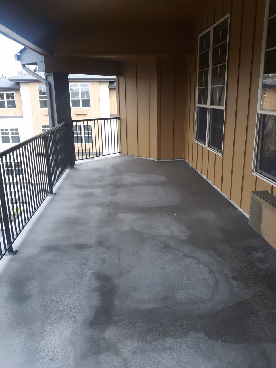 Covered outdoor balcony area with a concrete floor, metal railing, and beige siding walls with windows. The balcony overlooks another building with similar beige siding and multiple windows.