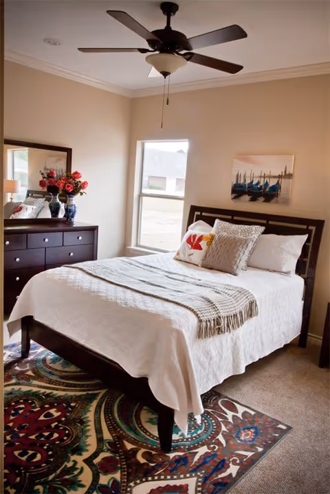 A cozy bedroom with a neatly made bed featuring white bedding, decorative pillows, and a beige throw blanket. There is a dark wooden dresser with a mirror on the left side, adorned with a vase of red flowers and other decorative items. A ceiling fan with a light fixture hangs above, and a window lets in natural light. A colorful patterned rug is placed under the bed, and a framed picture of boats on water hangs on the wall above the bed.
