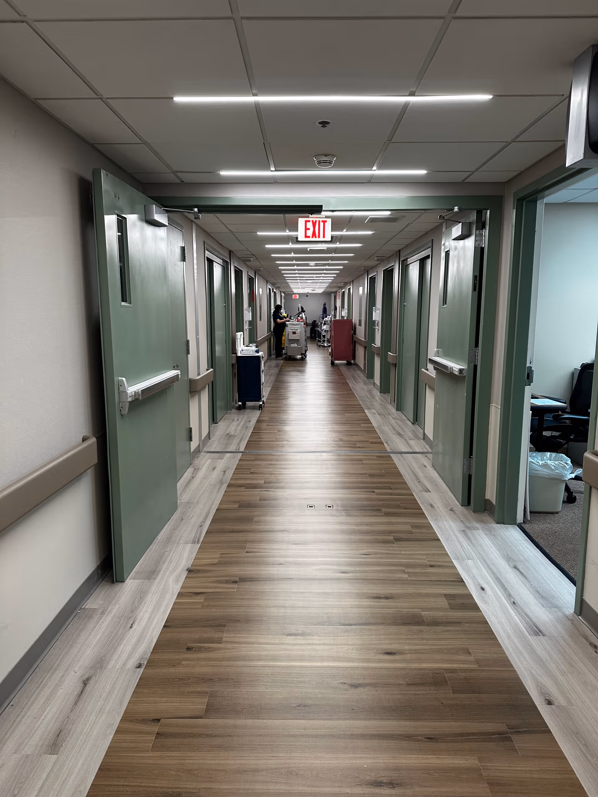 A long, well-lit hallway in a healthcare facility with green doors on both sides, some open. The floor has a wood-like finish with lighter borders. At the end of the hallway, a person is seen standing near medical carts. An illuminated exit sign hangs from the ceiling.