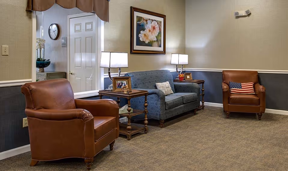 A cozy sitting area with two brown leather armchairs and a blue upholstered loveseat. Each side of the loveseat has a wooden side table with a lamp and framed photos. A painting of flowers hangs on the wall above the loveseat. The room has beige and dark blue walls with a carpeted floor. A mirror on the left wall reflects a door and a clock.