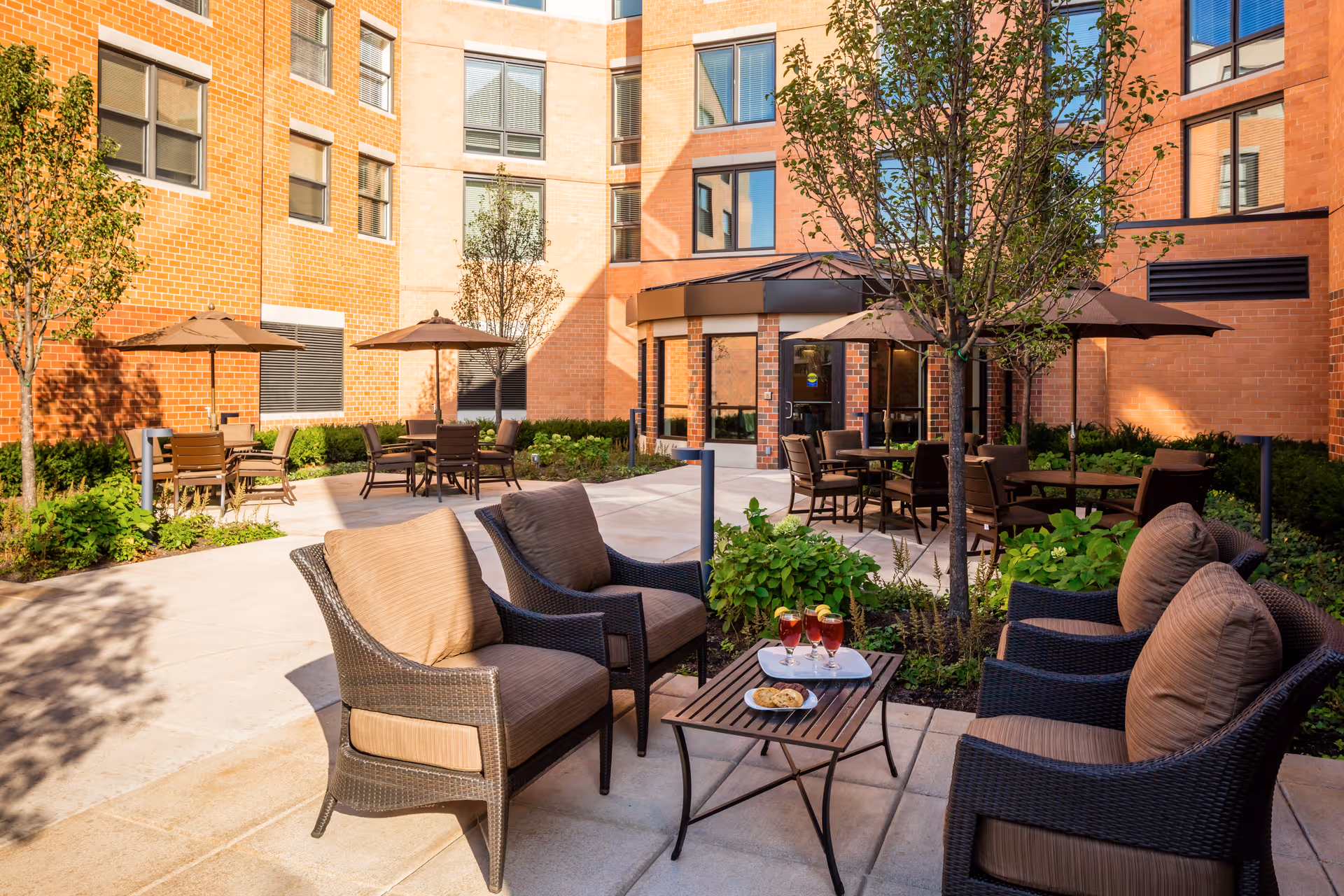 Courtyard patio with wicker lounge chairs, a small table, dining tables with umbrellas, and plants in front of a brick residential building.