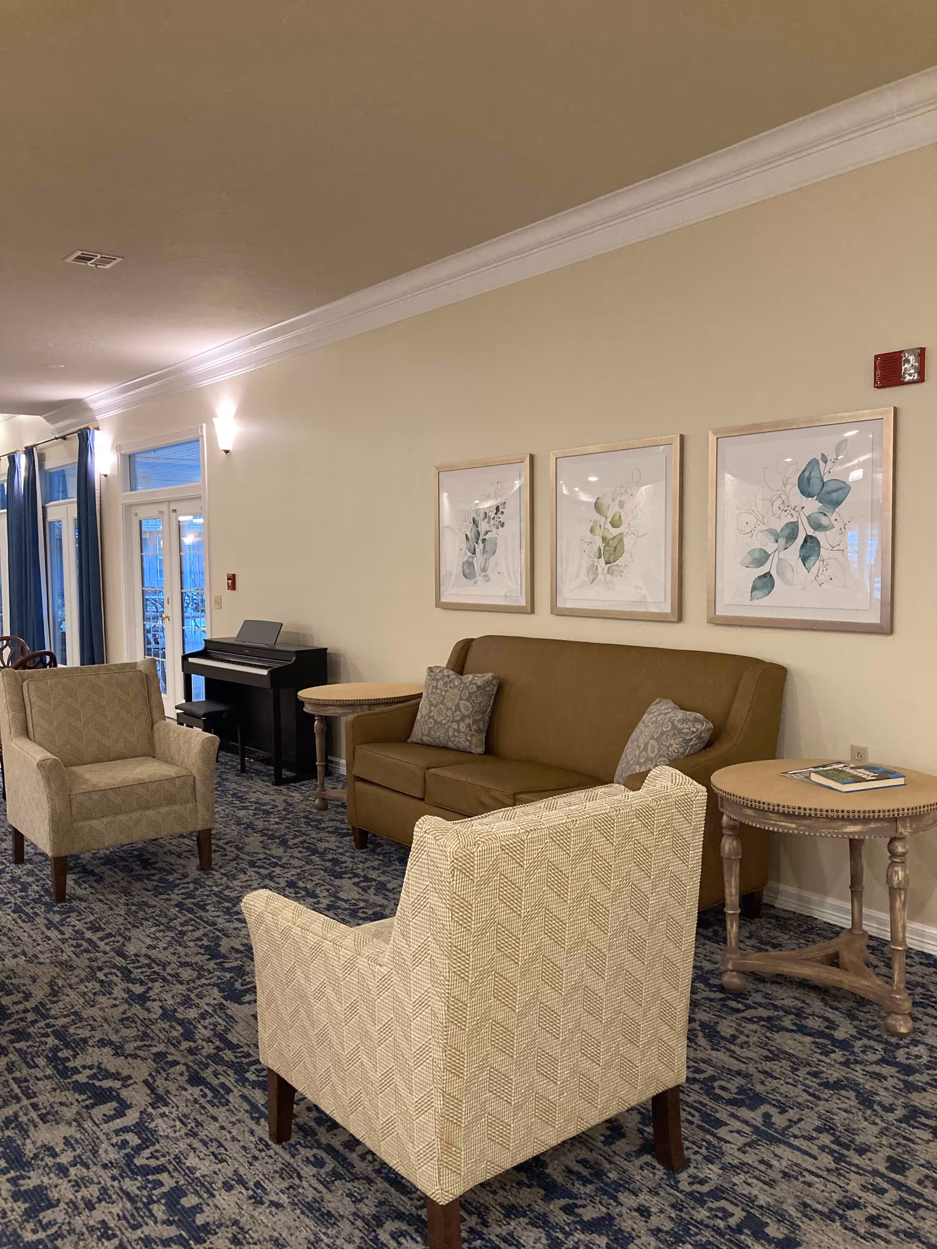 A cozy living room area in a senior living facility with two patterned armchairs and a brown sofa with two decorative pillows. There are three framed botanical prints on the wall above the sofa. A round wooden side table with a book on it is next to the sofa. In the background, there is a black piano and a set of glass doors with blue curtains. The room has a patterned blue carpet and cream-colored walls with crown molding.
