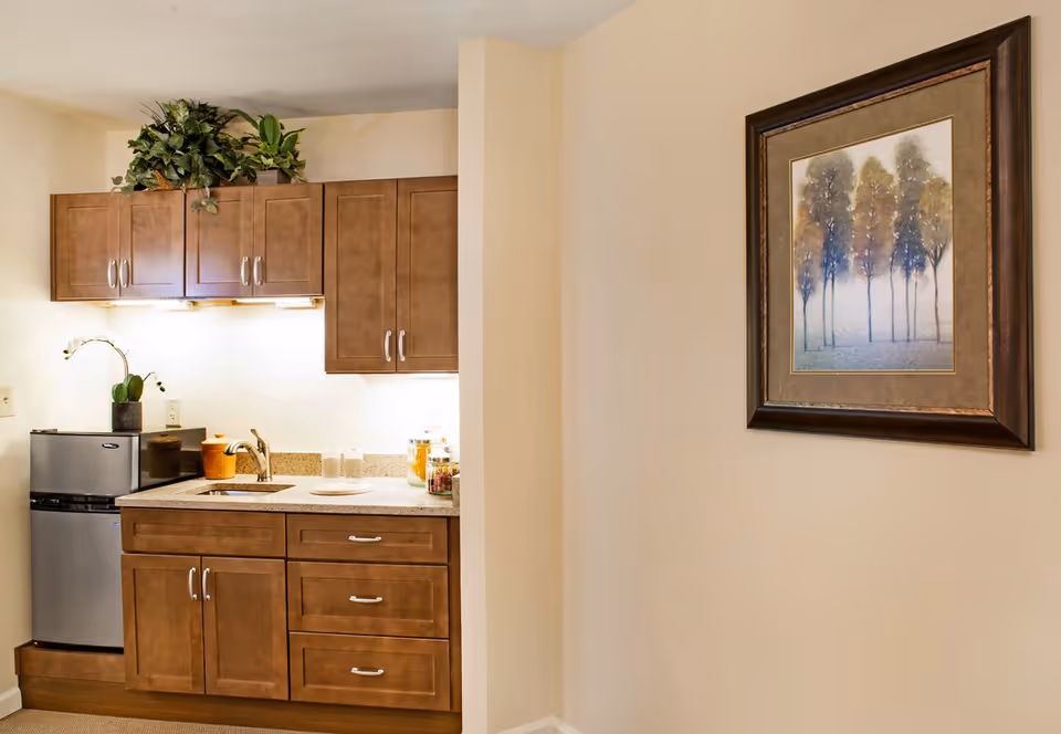Small kitchenette with wooden cabinets, a sink and mini fridge beside a wall with framed artwork.