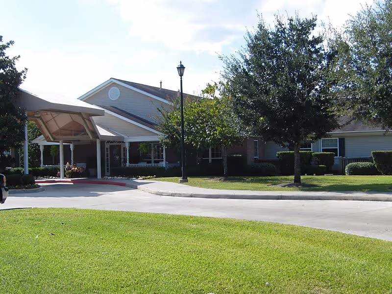Exterior view of a single-story senior living facility building with a covered entrance, surrounded by green grass, trees, and shrubs under a partly cloudy sky.
