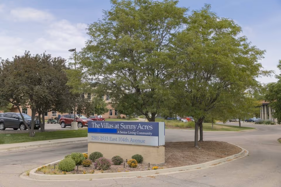 Outdoor view of the entrance sign for The Villas at Sunny Acres, a senior living community, located at 2501-2515 East 104th Avenue. The sign is surrounded by small shrubs and trees, with a parking lot and several cars visible in the background.