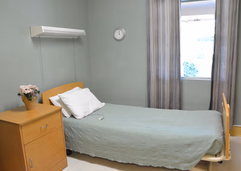 A simple bedroom in a rehabilitation center with a single bed covered in a light green blanket, two white pillows, a wooden nightstand with a vase of flowers, a wall clock, and a window with gray curtains letting in natural light.