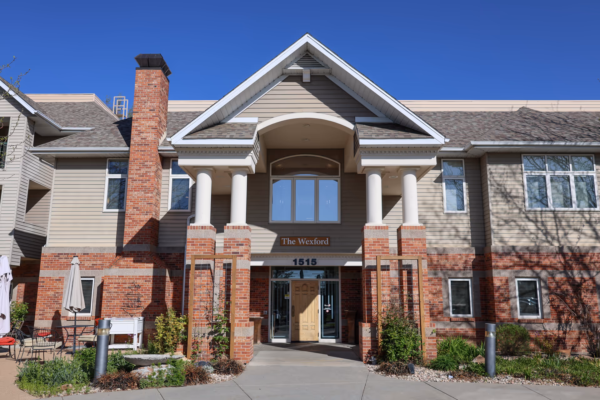 Front exterior view of The Wexford facility showing a two-story building with beige siding and red brick accents, white columns supporting a gabled roof over the entrance, and clear blue sky above.