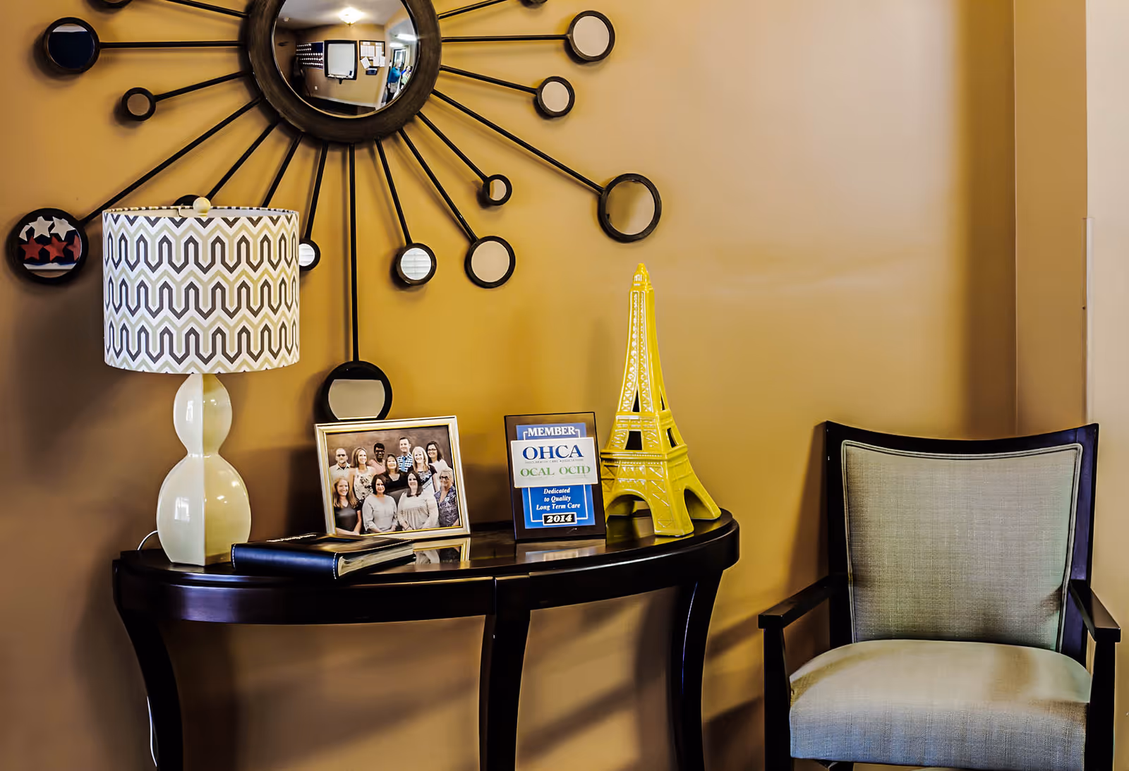 A small console table against a tan wall holds a patterned lamp, a framed group photo, a plaque and a yellow Eiffel Tower ornament beneath a decorative sunburst mirror, beside a cushioned armchair.
