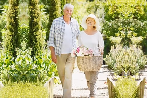 An elderly couple walking outdoors along a garden path surrounded by greenery. The man is wearing a plaid shirt over a white t-shirt and beige pants, while the woman is wearing a white hat, white top, and beige pants, carrying a basket filled with flowers. Both are smiling and enjoying the sunny day.