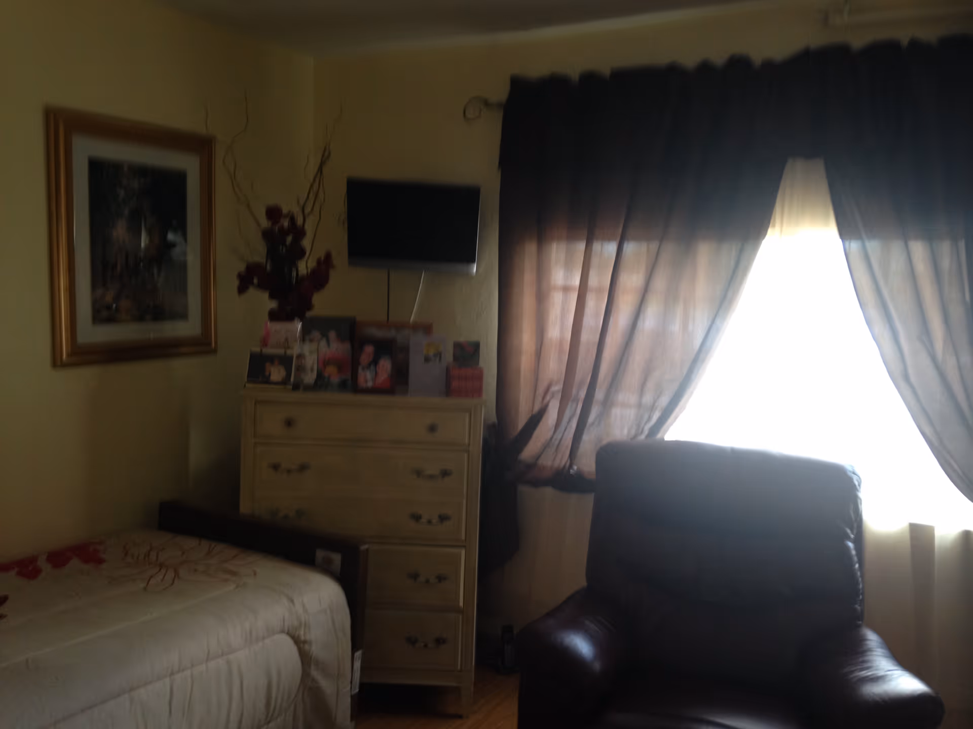 Small bedroom with a bed, dresser topped with photos, a wall-mounted TV, and a leather armchair by a curtained window.