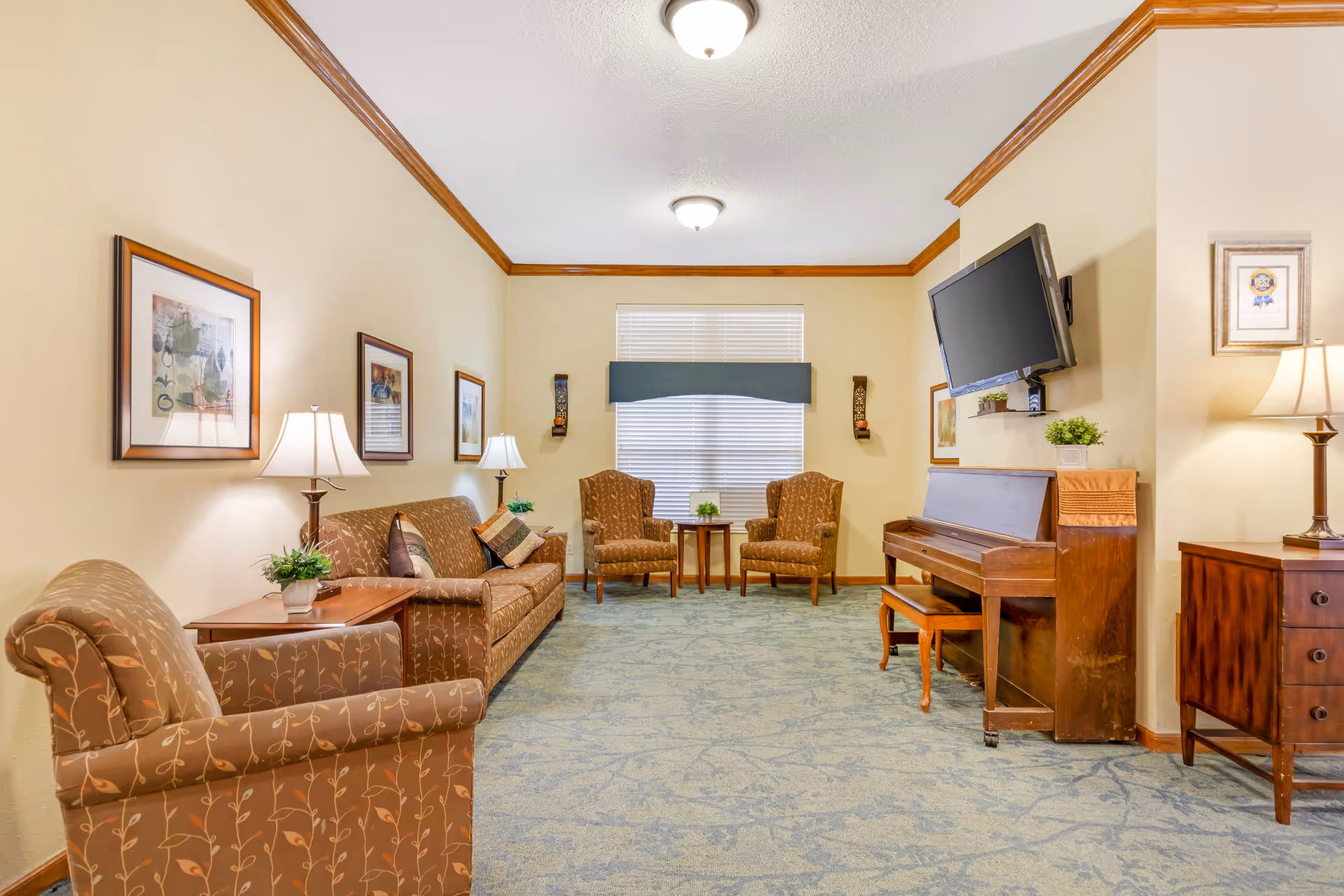 A cozy living room with beige walls and blue patterned carpet. The room features a brown patterned armchair and sofa with decorative pillows on the left side, two matching armchairs with a small wooden table between them in front of a window with blinds and a blue valance. On the right side, there is a wooden piano with a bench, a wall-mounted flat-screen TV above it, and a wooden side table with a lamp. The walls are decorated with framed artwork and sconces with candles.