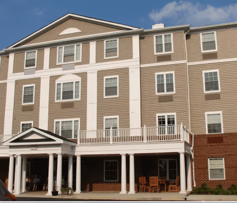 Exterior view of a three-story senior living facility building with beige and white siding, multiple windows, a covered entrance with white columns, and outdoor seating on the ground level under the balcony.