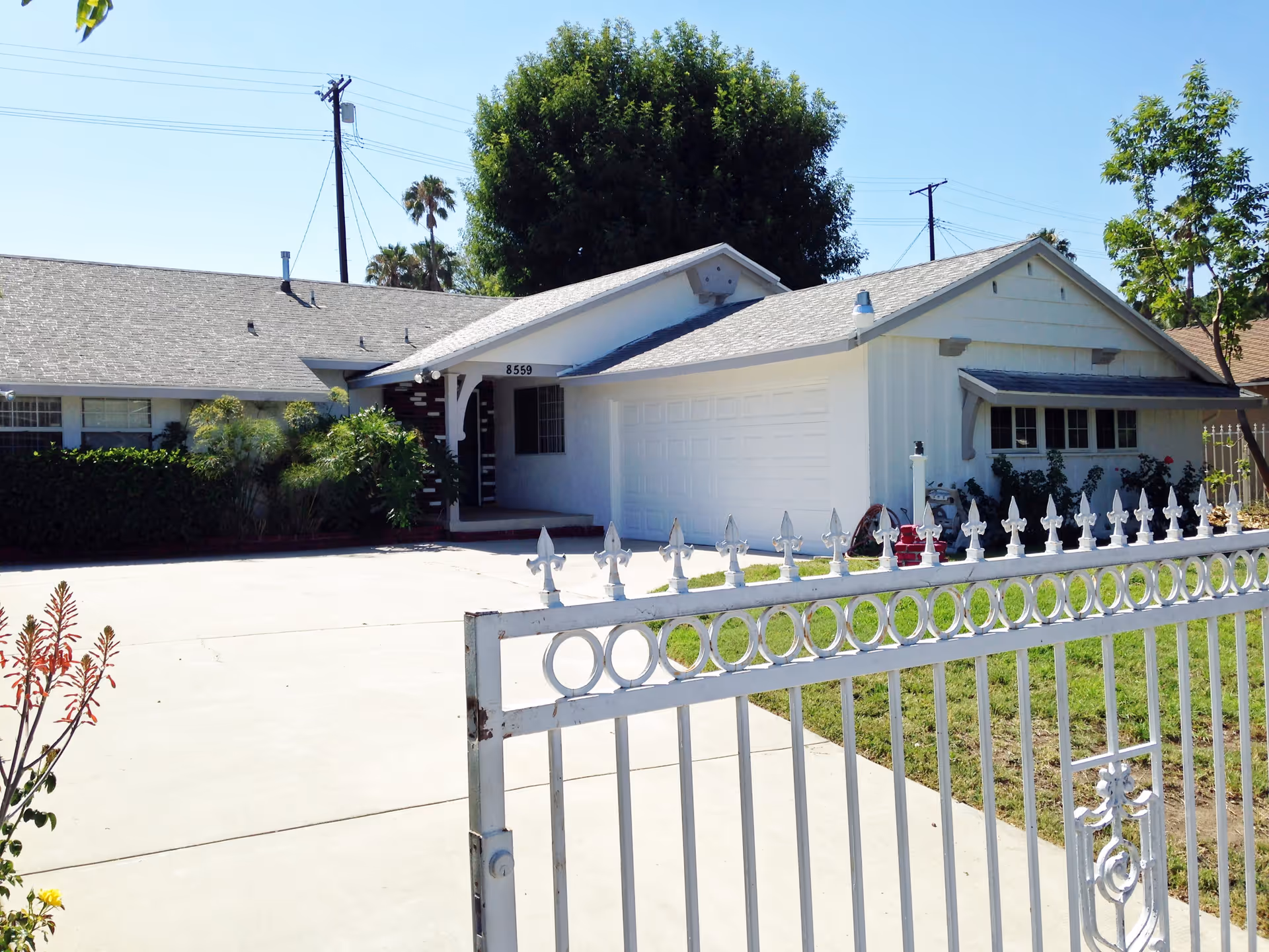 Front exterior view of a single-story residential building with a white garage door, a small porch, and a gated driveway. There are trees and bushes around the house, and a clear blue sky above.