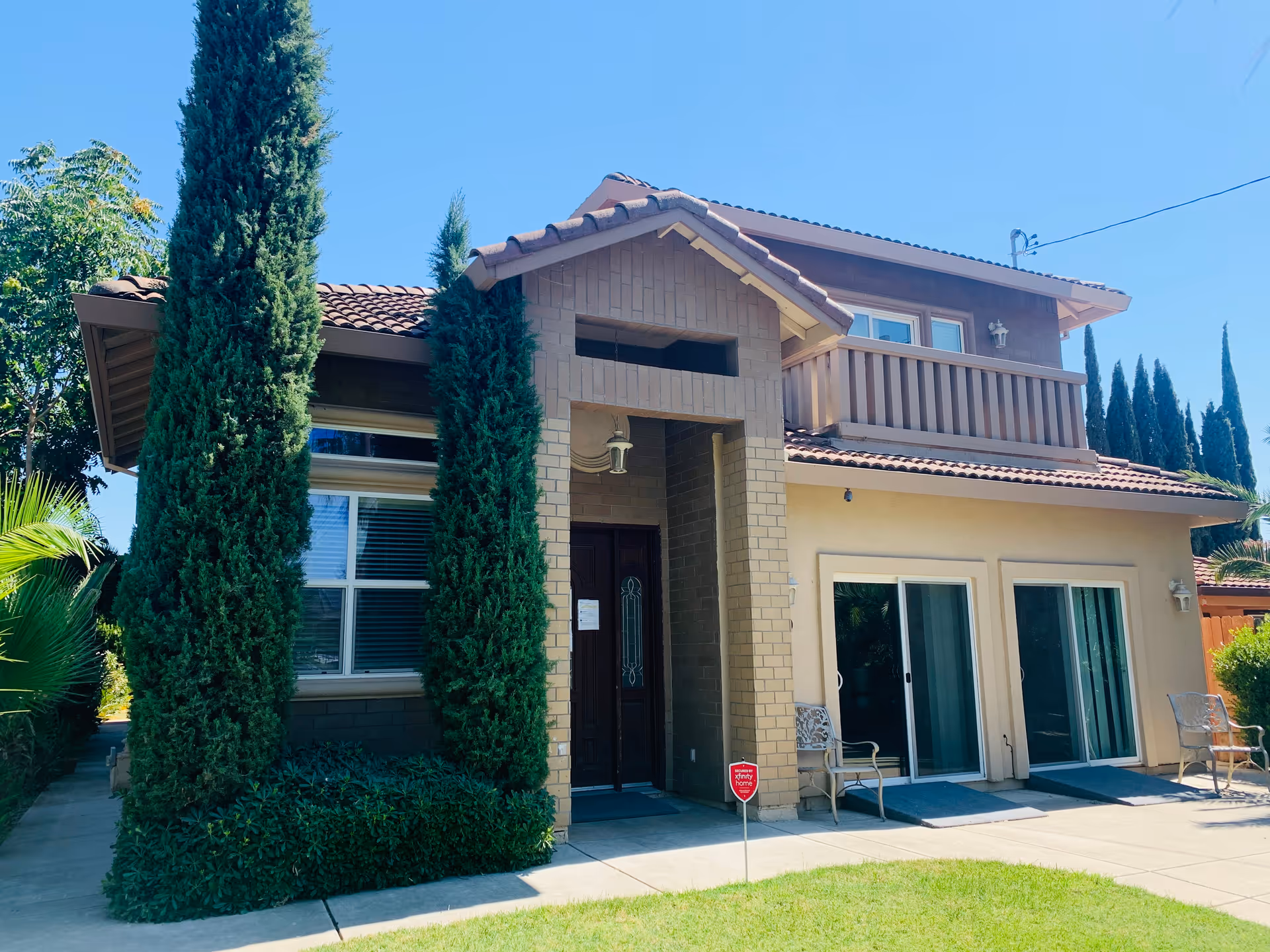 Front exterior view of a two-story residential building with a tiled roof, beige and brown walls, and a small balcony. The entrance is framed by two tall, narrow evergreen trees and a brick archway. There are two sliding glass doors on the right side with chairs placed outside. The lawn and sidewalk are visible in the foreground under a clear blue sky.