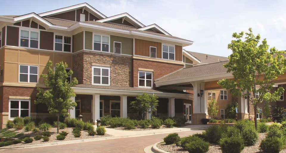 Front entrance of a multi-story senior living facility with a covered porte-cochère, windows, and landscaped grounds.