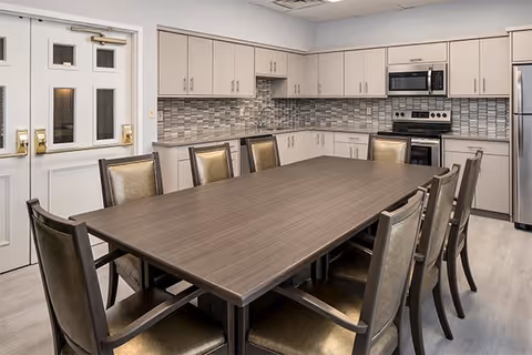 A modern kitchen and dining area with a large rectangular wooden table surrounded by eight chairs with cushioned seats. The kitchen features light-colored cabinets, a stainless steel refrigerator, microwave, and oven. The backsplash has a mosaic tile design in neutral tones. There are double doors with windows on the left side of the room.