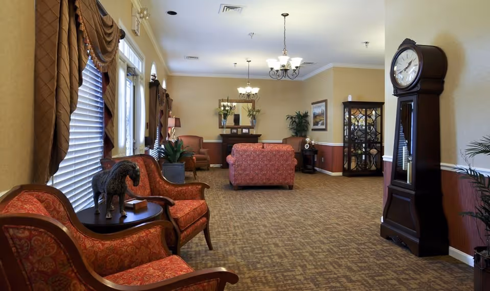 A spacious living room area with patterned carpet and beige walls. The room features multiple upholstered chairs and sofas in red and orange tones, a tall grandfather clock on the right, a glass display cabinet, and decorative plants. Large windows with blinds and brown drapes allow natural light to enter. Ceiling lights with chandeliers illuminate the room.