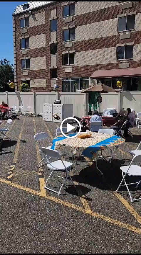 Outdoor patio area with several white folding chairs and tables covered with tablecloths. A few people are seated at the tables under a sunny sky. The area is adjacent to a multi-story brick building with windows and a white fence enclosing the space.