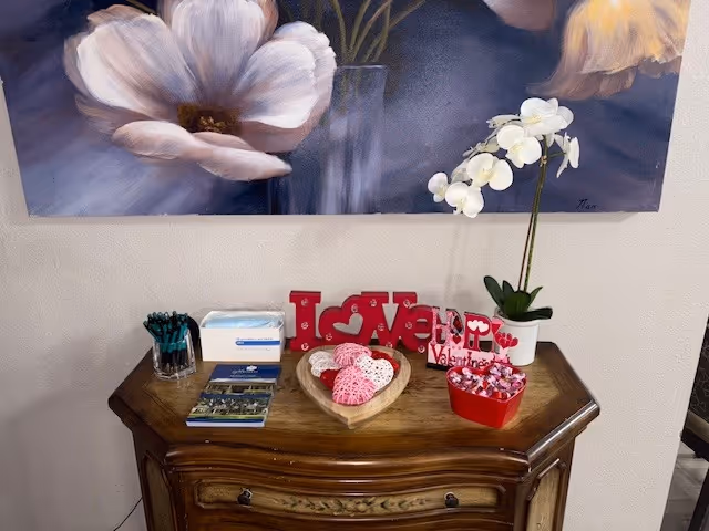 A wooden side table with Valentine-themed decorations including a red 'LOVE' sign, a heart-shaped basket with pink and white crocheted items, a red container with wrapped candies, a white orchid plant in a white pot, a box of tissues, a glass container with pens, and some brochures. Above the table is a large floral painting featuring white and pale pink flowers.