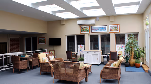 Sunlit communal lounge with wicker sofas and chairs, skylights, a TV and potted plants.
