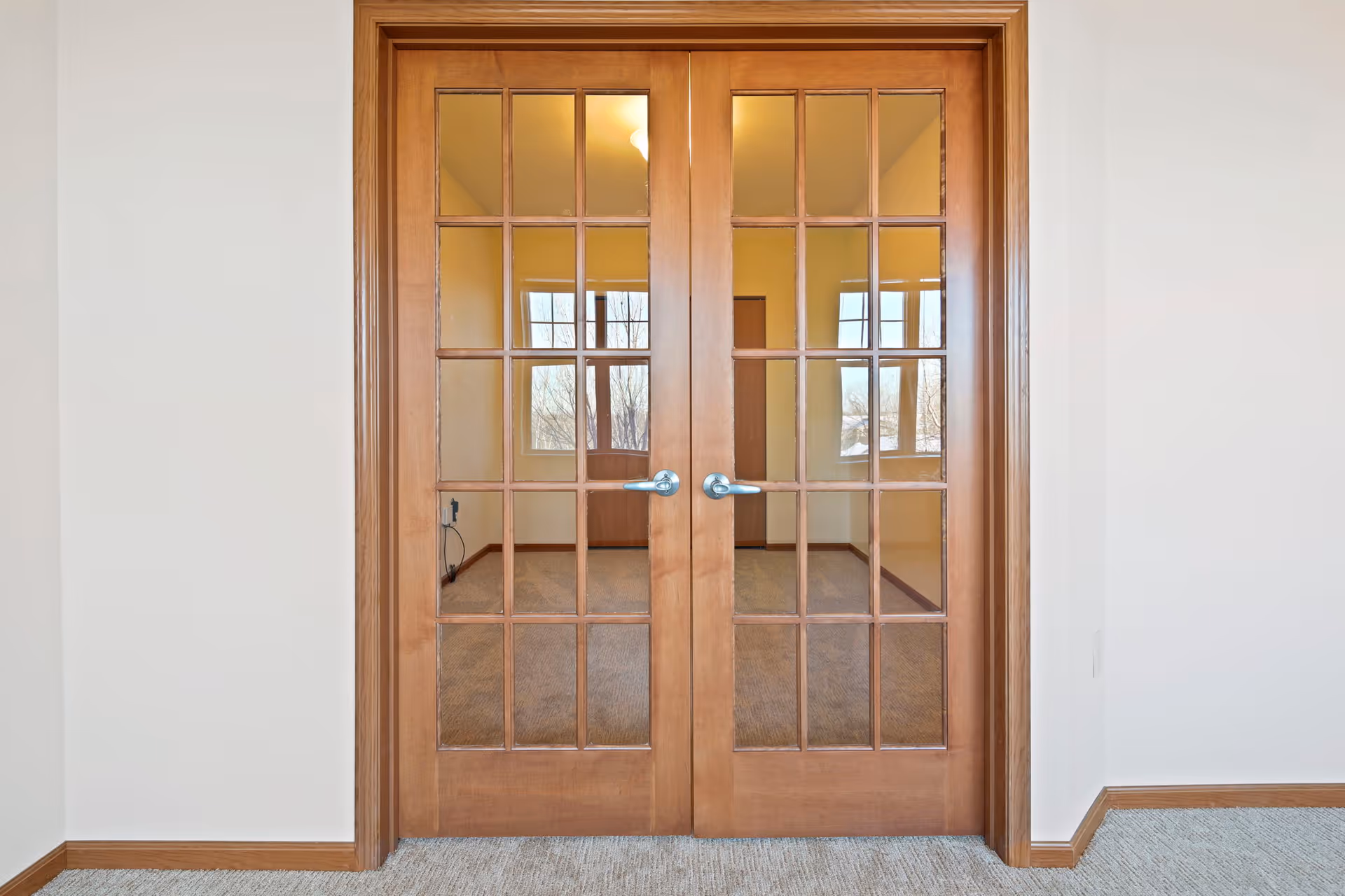 Closed wooden double doors with glass panes, showing a view into an empty room with carpeted floor, beige walls, and windows letting in natural light.