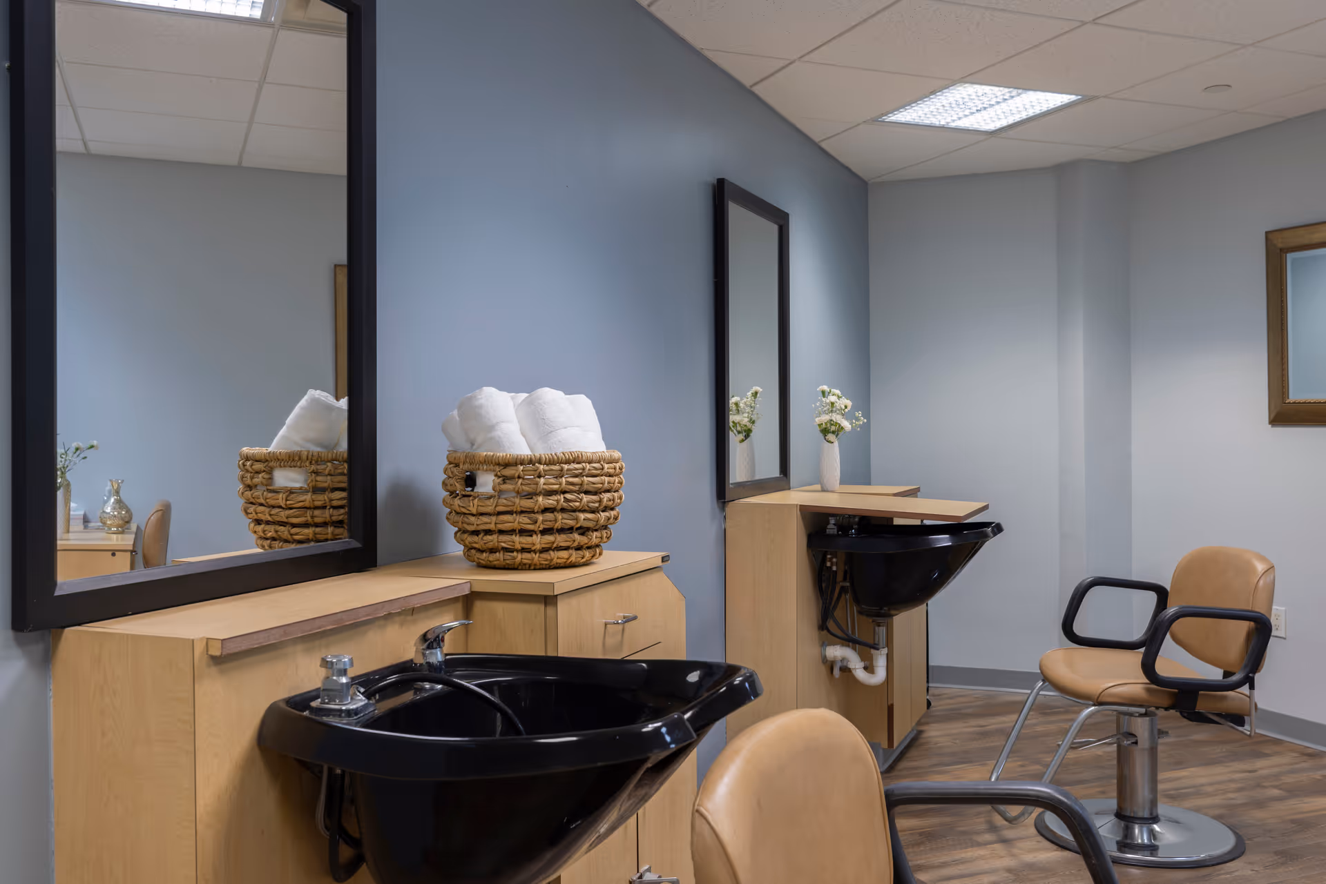 Interior of a salon area in an assisted living facility featuring two black hair washing sinks with wooden cabinets underneath, two large mirrors mounted on a blue wall, a basket with rolled white towels, a small vase with white flowers, and two tan salon chairs with black armrests on a wooden floor.