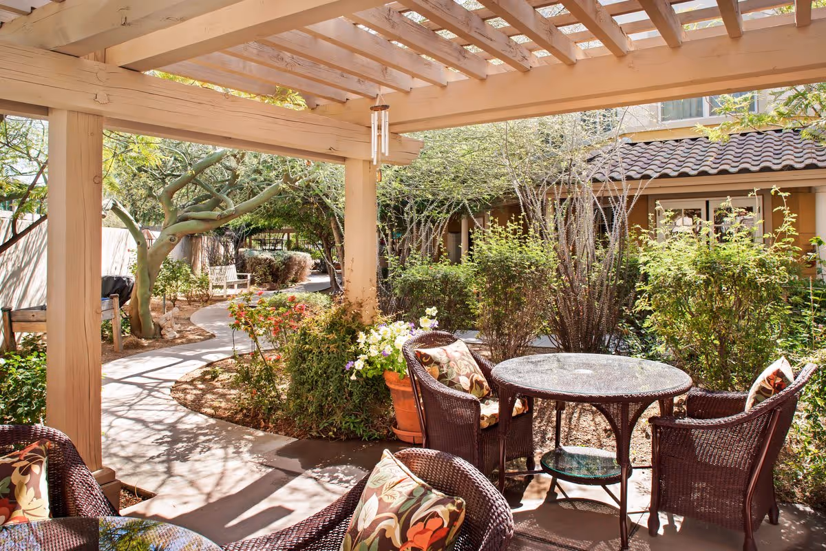 Outdoor patio area with a wooden pergola overhead, featuring a round glass-top table surrounded by four wicker chairs with floral cushions. The patio overlooks a garden with various bushes, flowering plants, and a winding concrete pathway leading to a bench under trees. The building with a tiled roof is visible in the background.