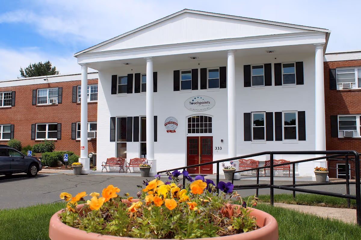 Front exterior view of Touchpoints at Manchester, a two-story building with white columns, red doors, and black window shutters. There are flower pots with colorful flowers and benches near the entrance, along with a ramp for accessibility.
