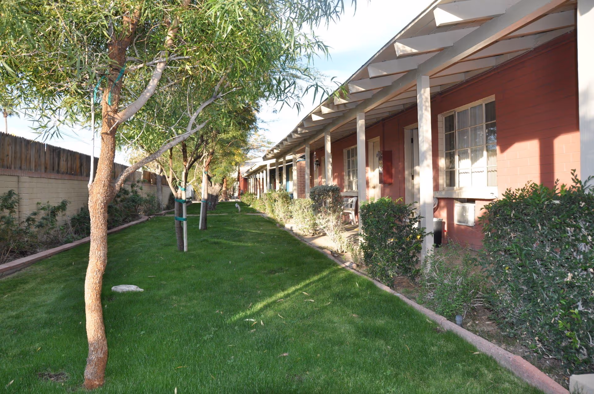 A grassy outdoor area with a row of small trees and bushes alongside a building with a covered porch. The building has red walls, white pillars, and several windows and doors. A wooden fence runs along the left side of the grassy area.