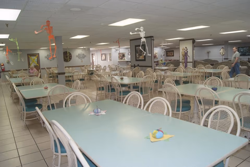 A large dining room with multiple light blue tables and white chairs with blue cushions. The room is decorated with hanging skeleton decorations and small decorative pumpkins on the tables. There are a few people visible in the background, and the room has a tiled floor and fluorescent ceiling lights.