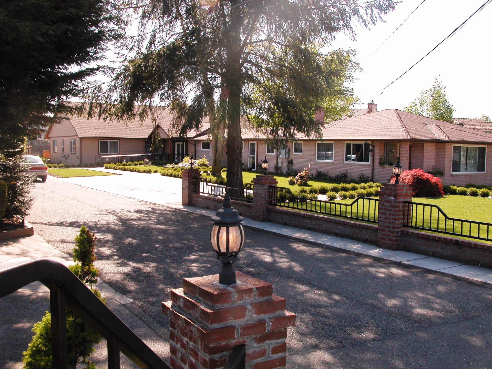 Exterior view of a single-story residential building with a well-maintained lawn, brick pillars with lanterns, and a paved driveway. Trees and shrubs surround the property under a sunny sky.