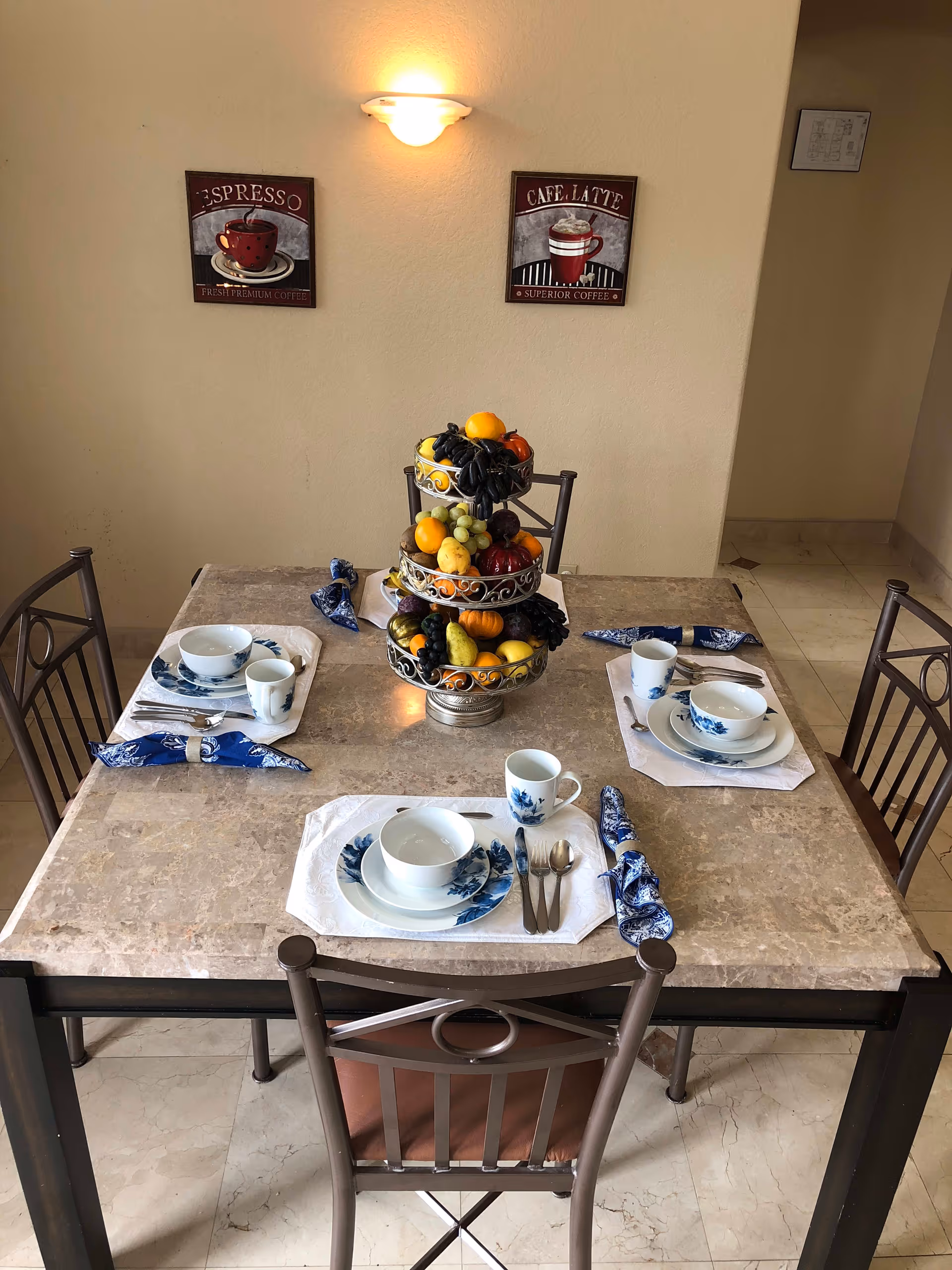 A dining table set for three with blue and white dishware, silverware, and blue patterned napkins. A three-tiered fruit stand with various fruits is placed in the center of the table. The room has beige walls with two coffee-themed wall decorations and a wall light above the table.