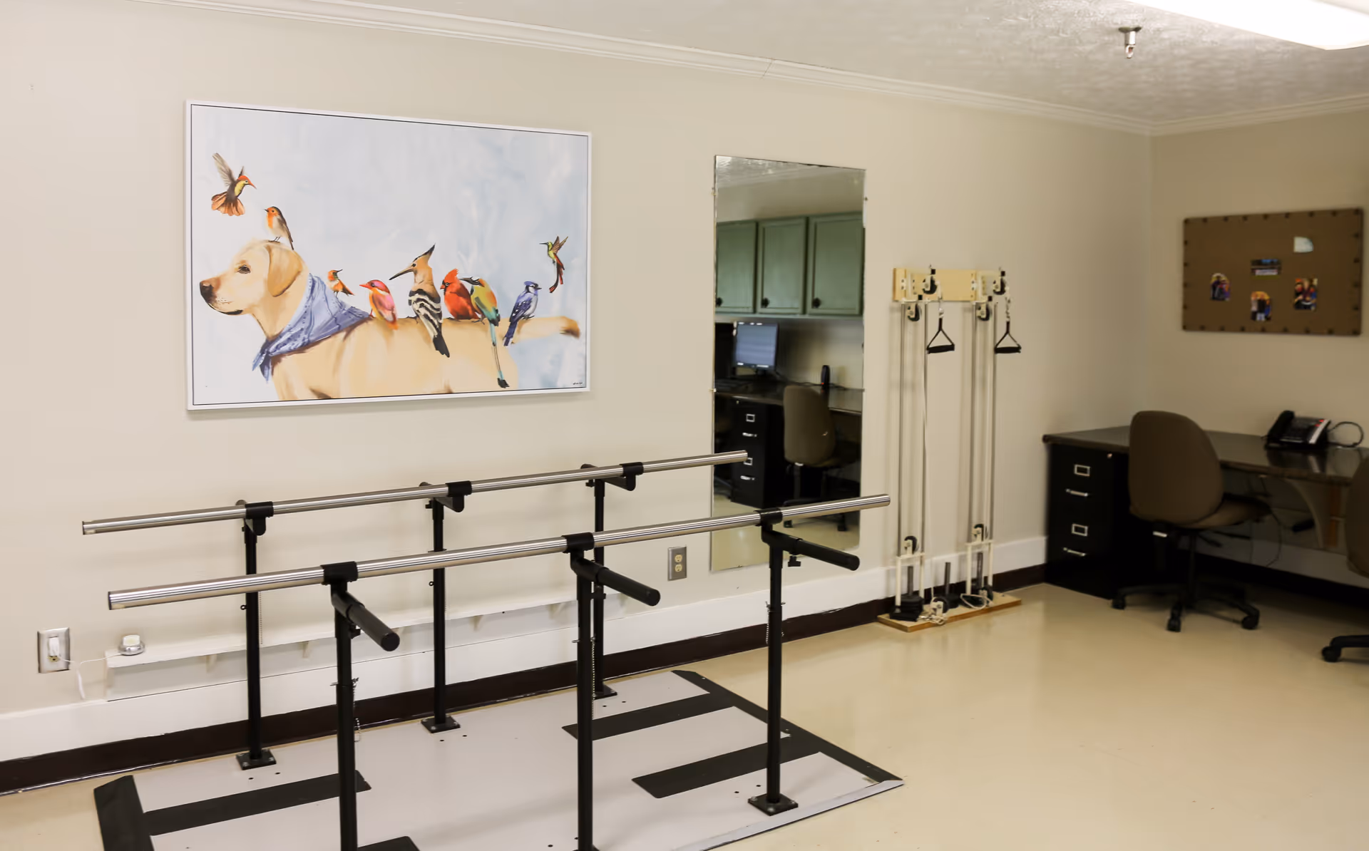 A rehabilitation room with parallel walking bars, a desk and chairs, a wall mirror and a colorful dog-and-birds painting.