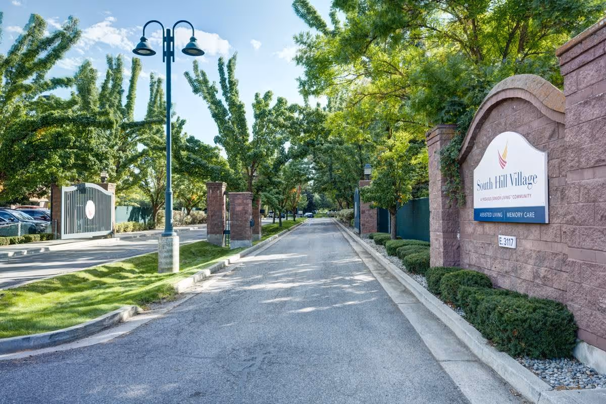 Entrance road to South Hill Village senior living community lined with trees and green landscaping, featuring a brick wall with the community's sign on the right side and a gated entrance on the left.