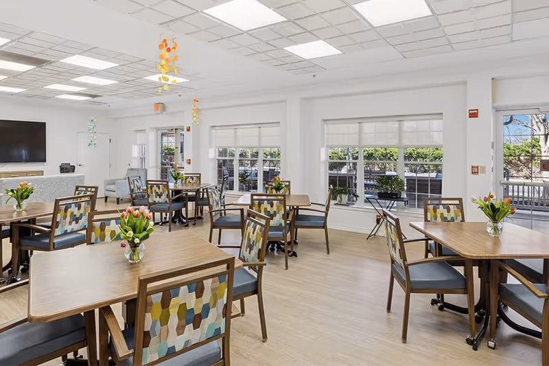 A bright and spacious dining room in a senior living facility with several wooden tables and chairs featuring colorful patterned backs. Each table has a small vase with fresh flowers. Large windows allow natural light to fill the room, and there is a door leading to an outdoor area. The ceiling has recessed lighting and decorative hanging ornaments.