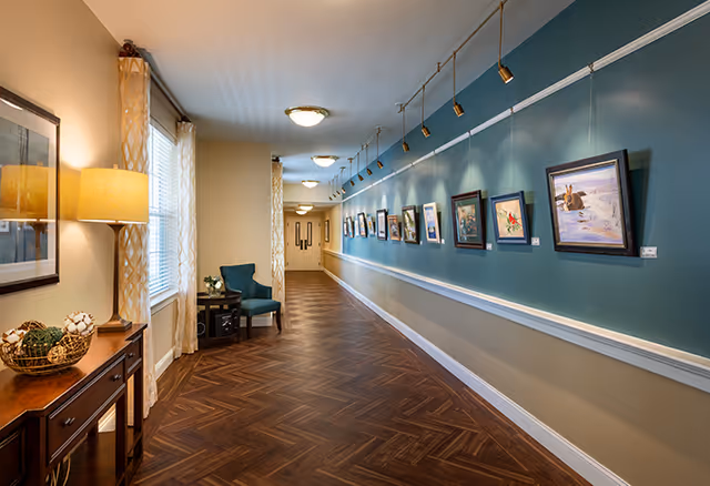 A well-lit hallway in a senior living facility with wooden herringbone patterned flooring and a teal accent wall displaying framed artwork. On the left side, there is a wooden console table with a lamp and decorative items, next to a window with patterned curtains and a blue upholstered chair beside a small table with a plant.