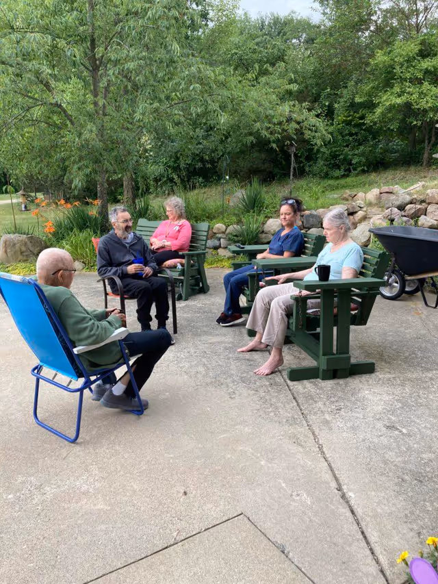 Five people sitting outside on a concrete patio surrounded by greenery and rocks. Four of them are seated on green wooden chairs, and one person is seated on a blue folding chair. They appear to be engaged in conversation or relaxing, with trees and plants in the background.