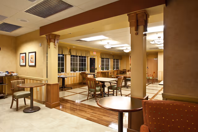 Interior view of a dining area in a senior living facility with multiple wooden tables and chairs arranged neatly. The room features warm beige walls, large windows with valances, and ceiling lights providing soft illumination. There are decorative columns and framed artwork on the walls, creating a cozy and inviting atmosphere.