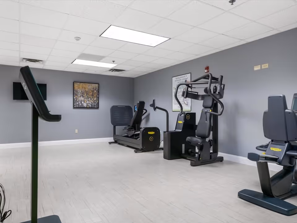 A fitness room with exercise equipment including a treadmill, a recumbent bike, and a multi-function strength training machine. The room has gray walls, a light-colored floor, a ceiling with fluorescent lights, a wall-mounted TV, and framed artwork on the wall.