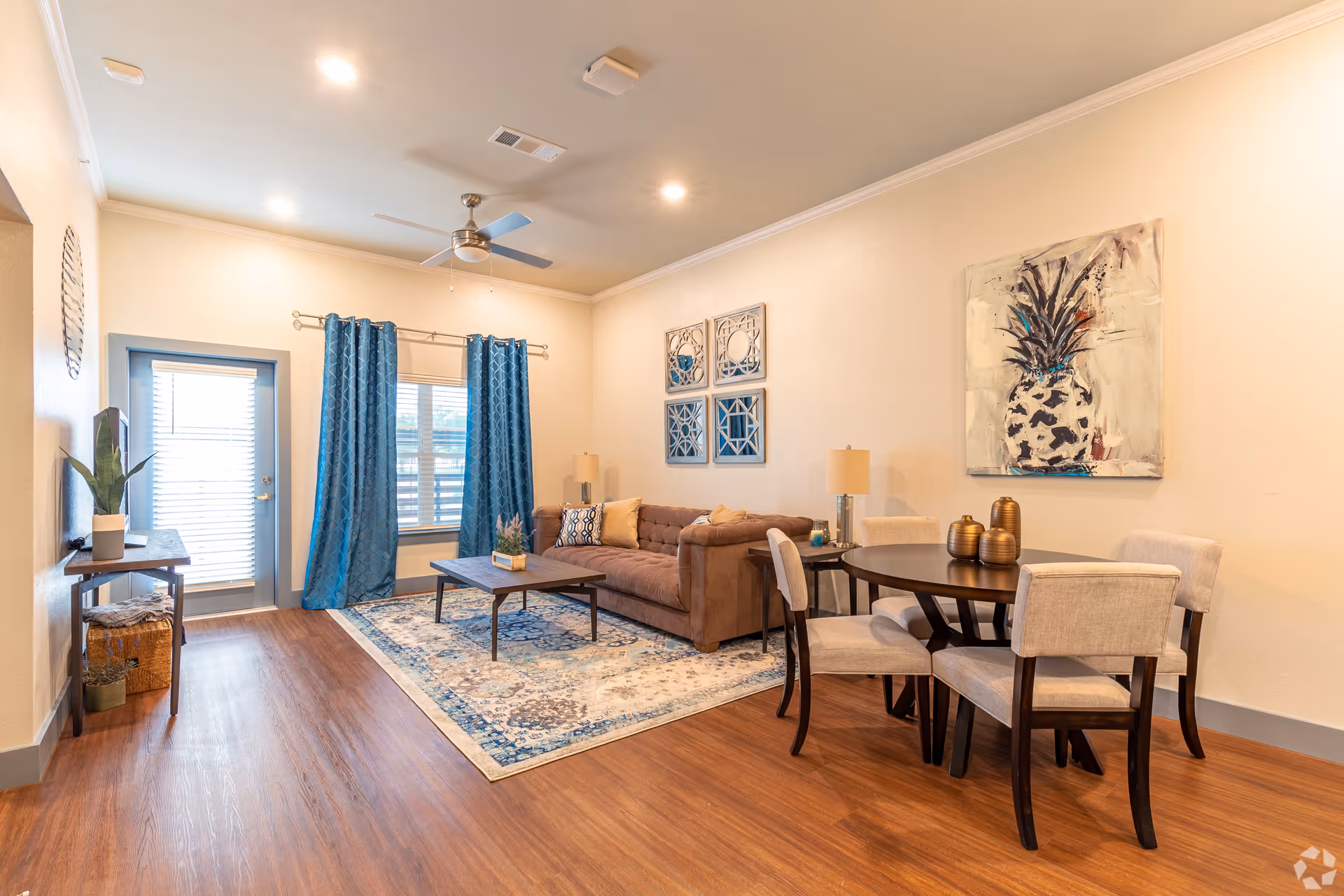 Bright open living area with a sofa, coffee table and rug, blue curtains, and a round dining table with four chairs.
