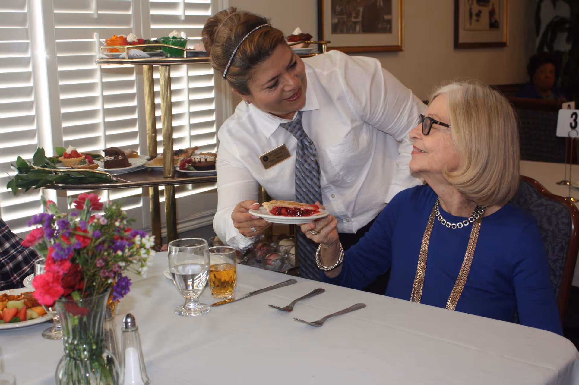A dining room scene where a staff member in a white shirt and tie is serving a slice of pie to an elderly woman wearing glasses and a blue top. The table is set with glasses of water and juice, silverware, and a vase of colorful flowers. A tiered tray with various desserts is visible in the background.