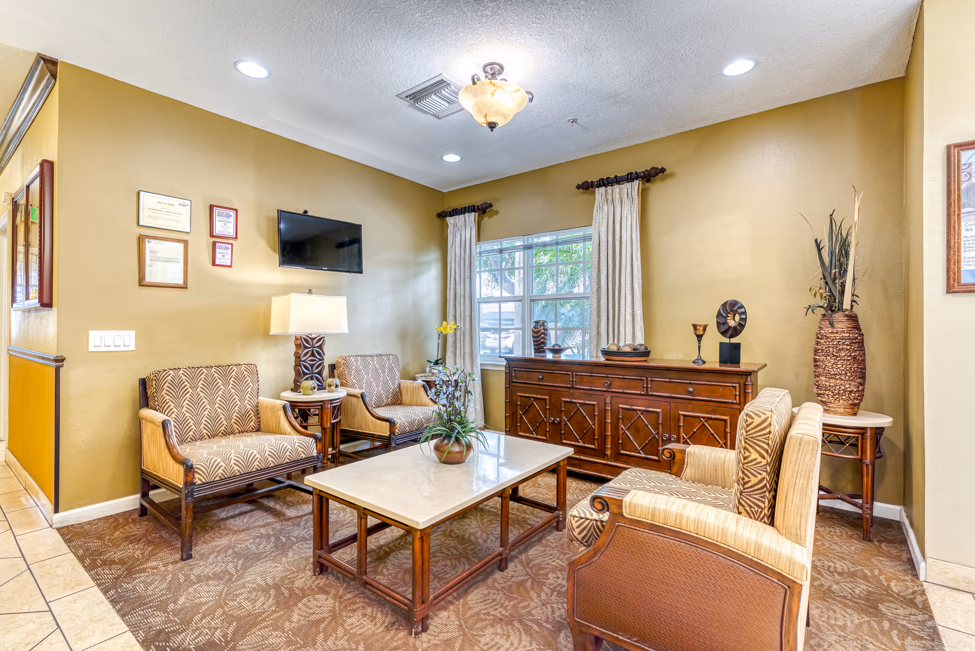 A cozy living room area with four patterned armchairs arranged around a rectangular coffee table with a plant centerpiece. There is a wooden sideboard against the wall with decorative items on top, a window with curtains, a wall-mounted TV, and framed certificates on the wall. The room has warm beige walls and a carpeted floor.