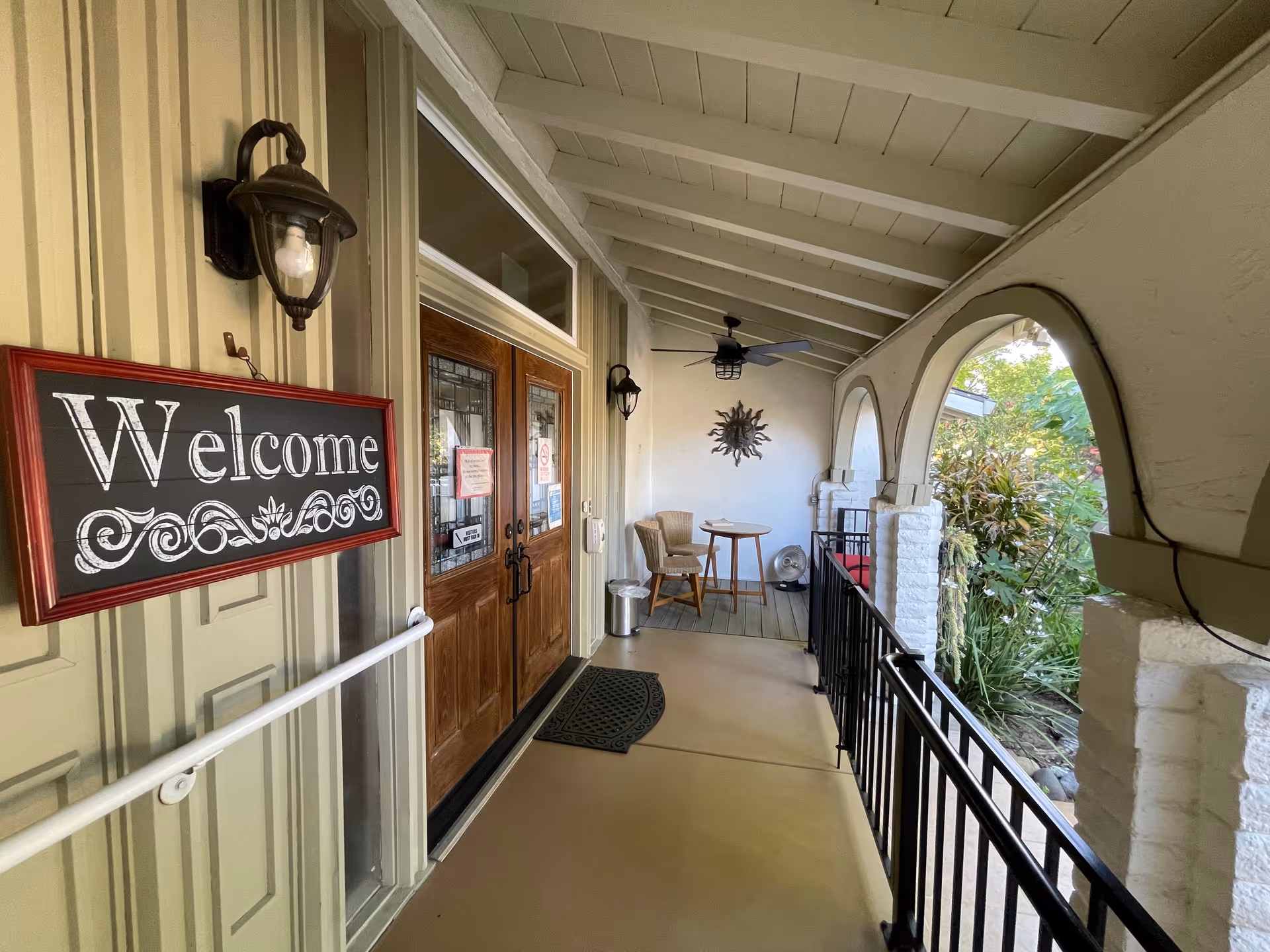 Covered porch area with a 'Welcome' sign on the left wall, two wooden doors with glass panels, a small table with two chairs, a ceiling fan, and arched openings showing greenery outside.