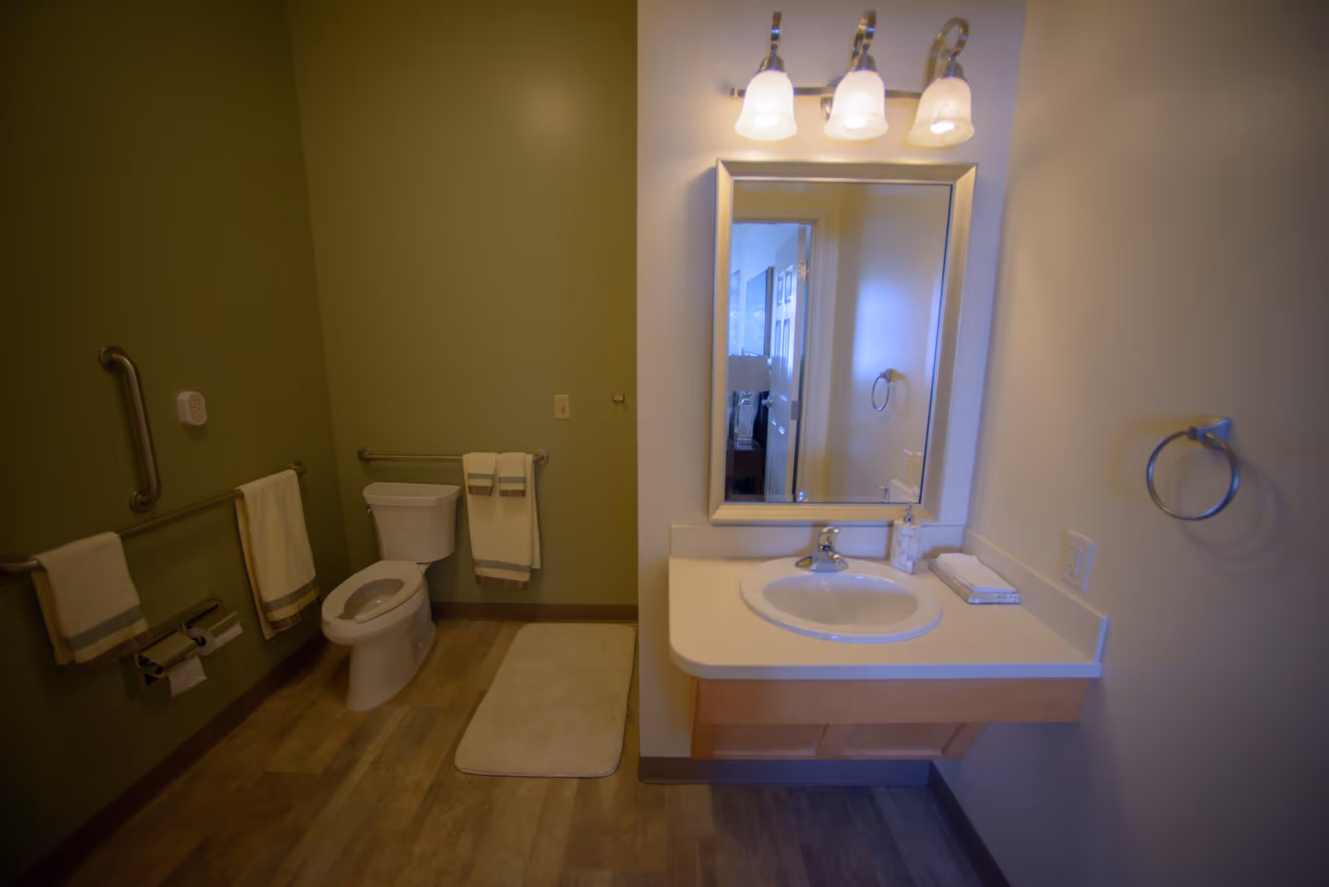 A clean bathroom with a toilet, a wall-mounted sink with a mirror above it, and three light fixtures. The walls are painted green and white, with grab bars and towel racks holding white towels. There is a beige bath mat on the wooden floor.