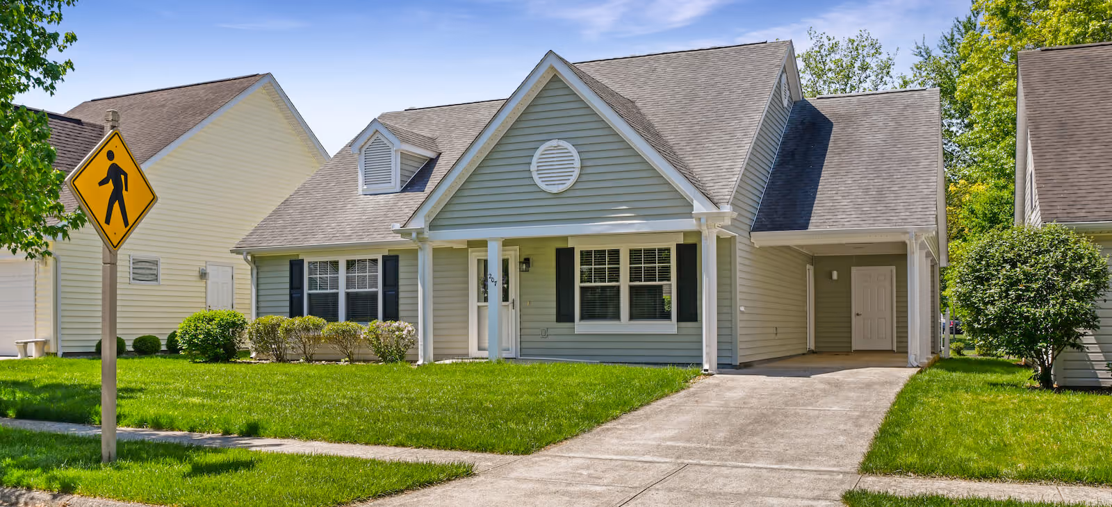 A single-story residential house with light gray siding and white trim, featuring a front porch with columns, black shutters on the windows, a driveway leading to a covered carport, and a well-maintained green lawn with bushes. A pedestrian crossing sign is visible on the left side near the sidewalk.