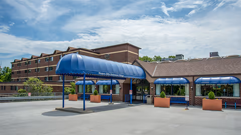 Front entrance of a brick senior living facility with multiple blue awnings covering the drop-off driveway.