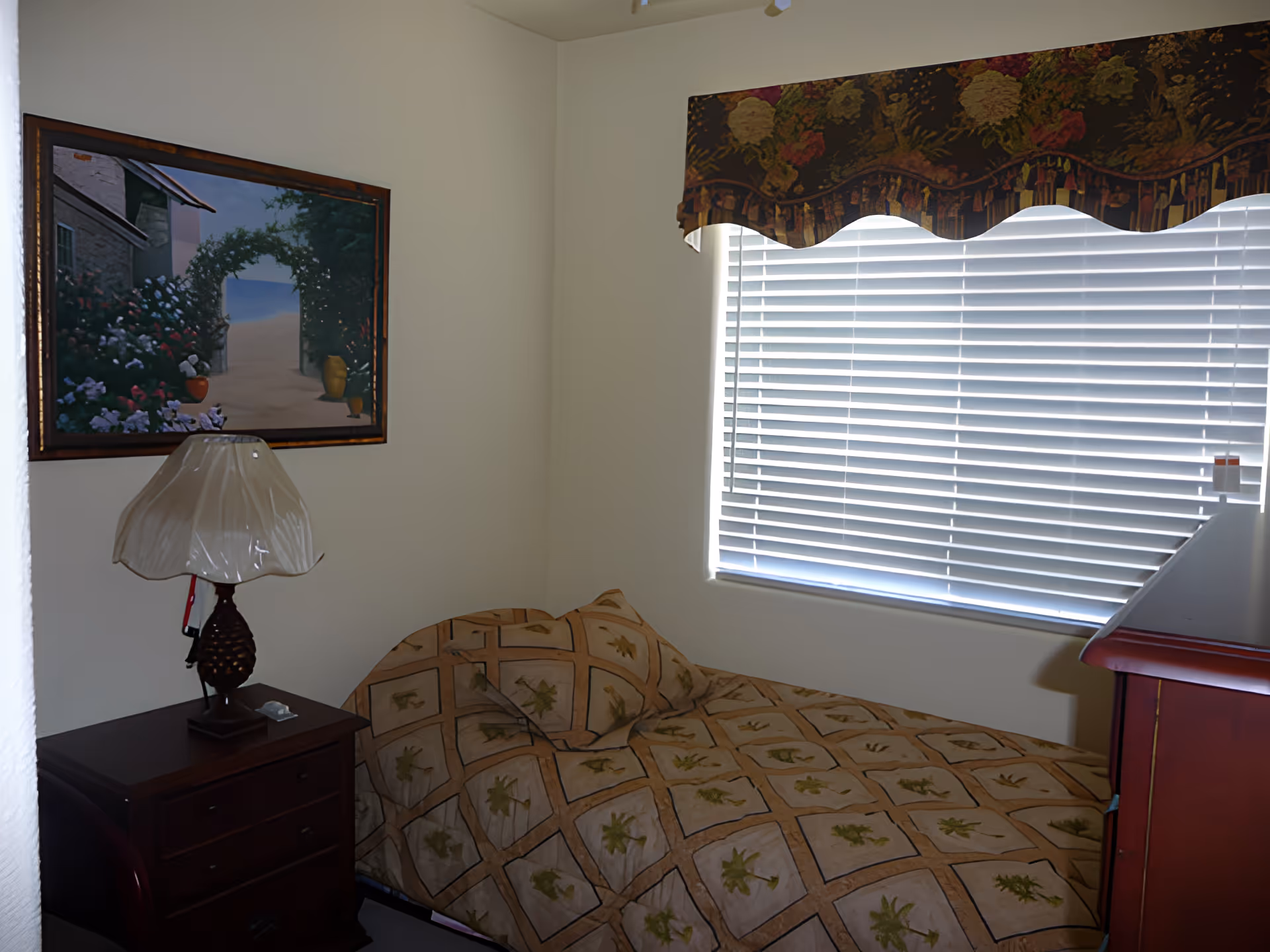 A small bedroom with a single bed covered in a beige quilt with leaf patterns. Next to the bed is a dark wooden nightstand with a lamp that has a translucent beige shade. On the wall above the nightstand is a framed painting depicting a garden archway with flowers. A window with white blinds and a floral valance is on the right side of the room.