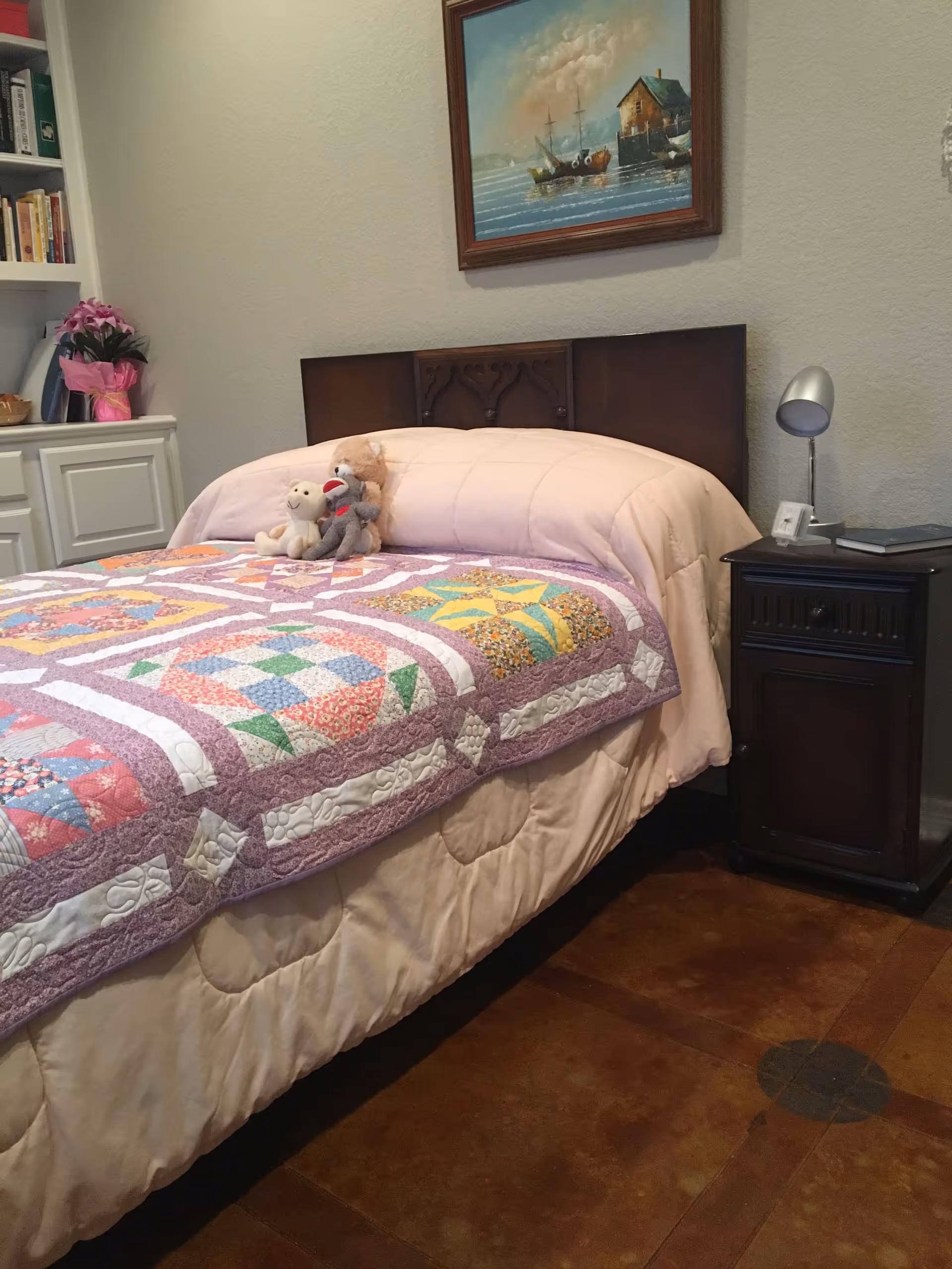 A cozy bedroom with a bed covered in a colorful patchwork quilt and a light pink comforter. Three stuffed animals are placed on the bed near the headboard. To the right of the bed is a dark wooden nightstand with a small lamp and a clock. On the wall above the bed hangs a framed painting of boats on water near a house. To the left, there is a white built-in cabinet with books and a pink flower arrangement on top.