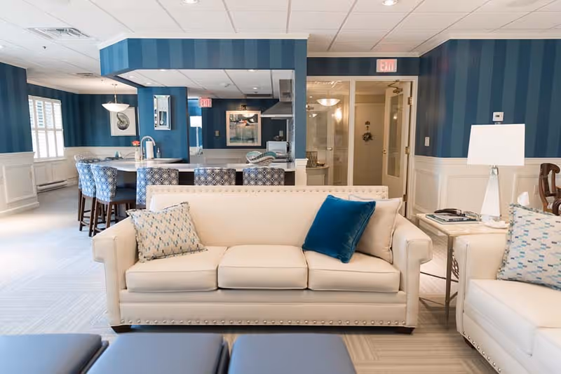 A bright and spacious living room area with cream-colored sofas adorned with patterned and solid blue throw pillows. Behind the sofas is a kitchen area with a breakfast bar and four patterned bar stools. The walls are decorated with blue striped wallpaper and white wainscoting. The room has a modern, clean look with a mix of blue and neutral tones, and there is a lamp on a side table next to one of the sofas.
