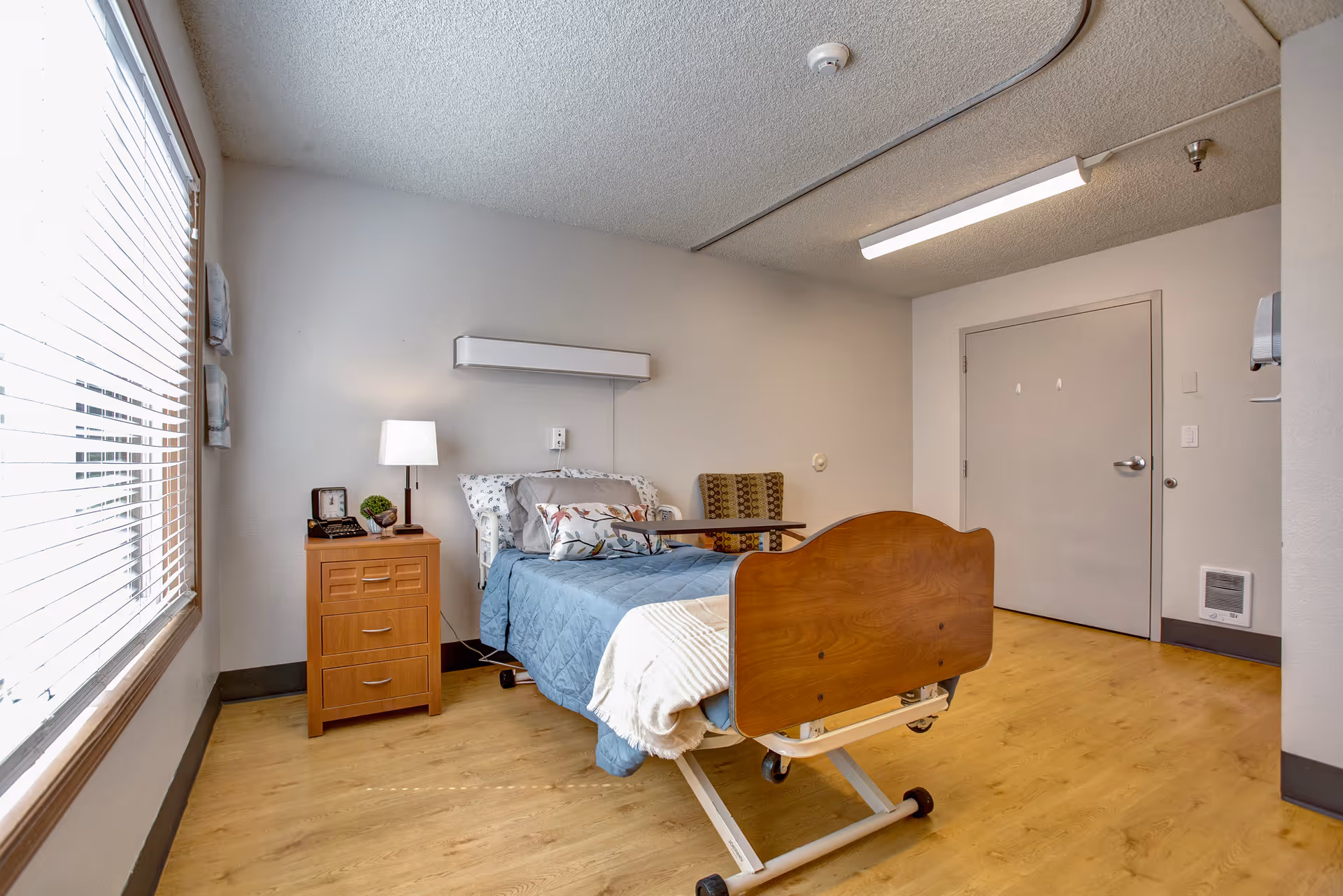 Private resident bedroom with a hospital-style bed, nightstand, lamp and large window in a senior living facility.